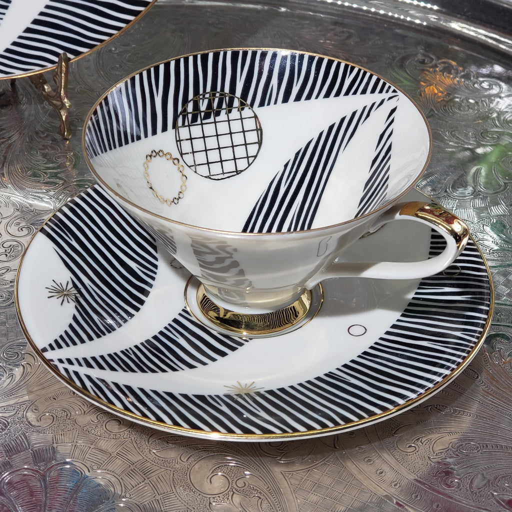 A white teacup with black and gold stripes and a matching saucer sits on a patterned tablecloth.