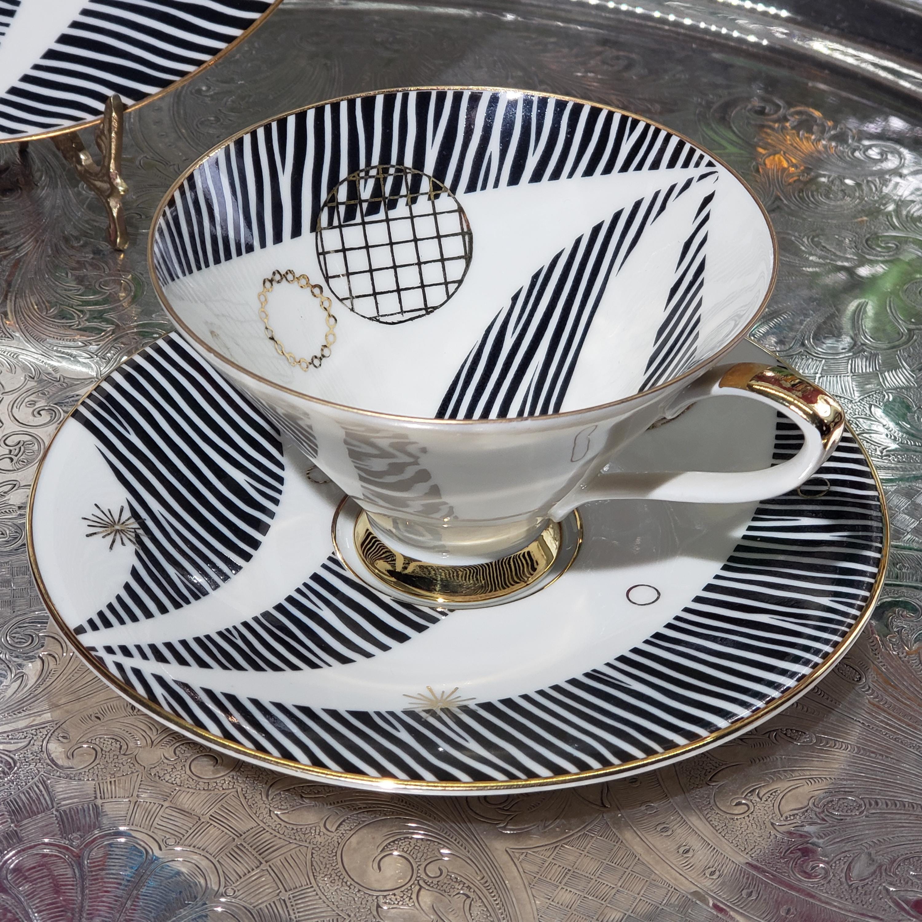 A white teacup with black and gold stripes and a matching saucer sits on a patterned tablecloth.