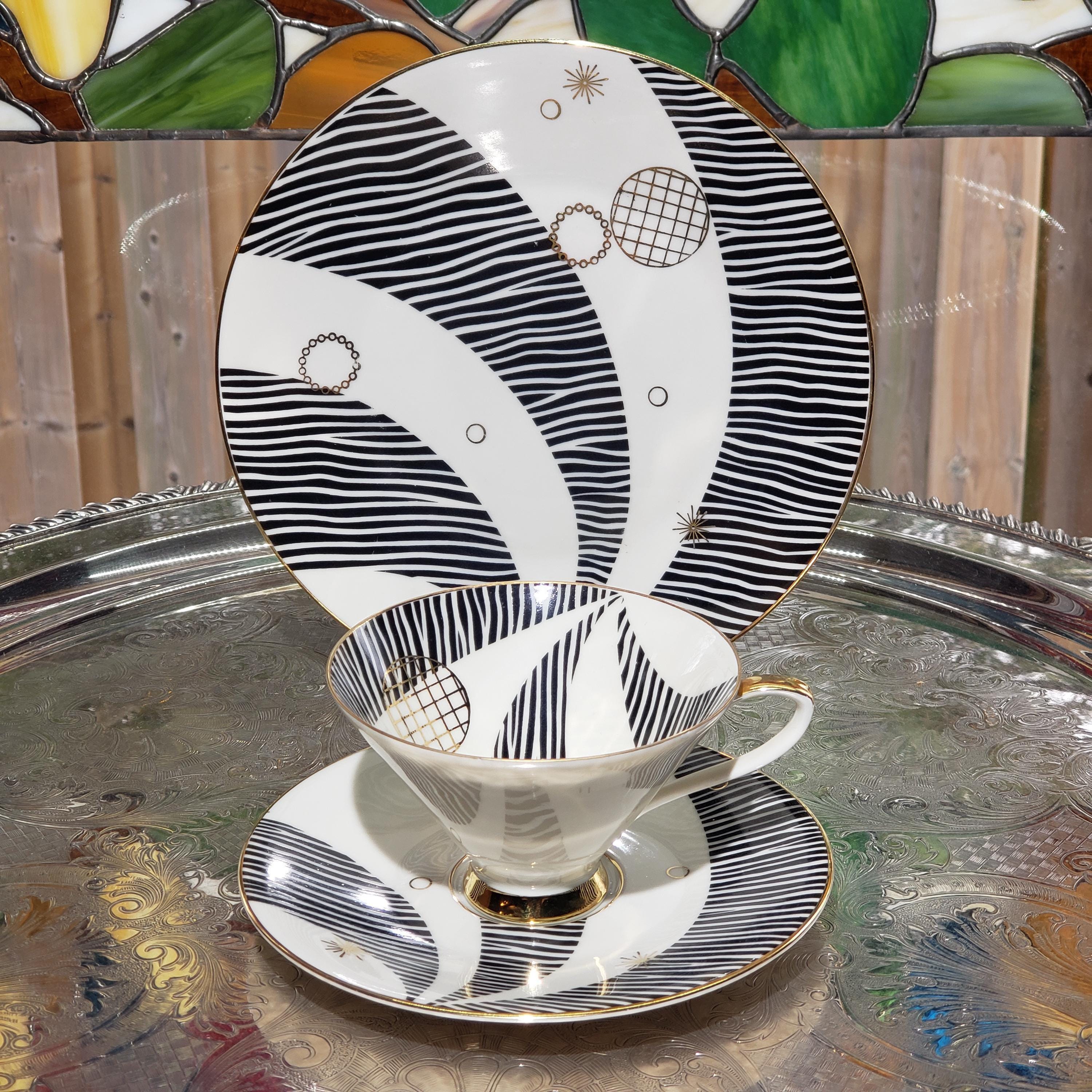 A white and black striped plate with gold accents and a matching cup are displayed on a glass table, with a stained glass window in the background.