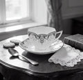 A black and white image of a tea set on a wooden table, including a teacup, saucer, and a lace doily.