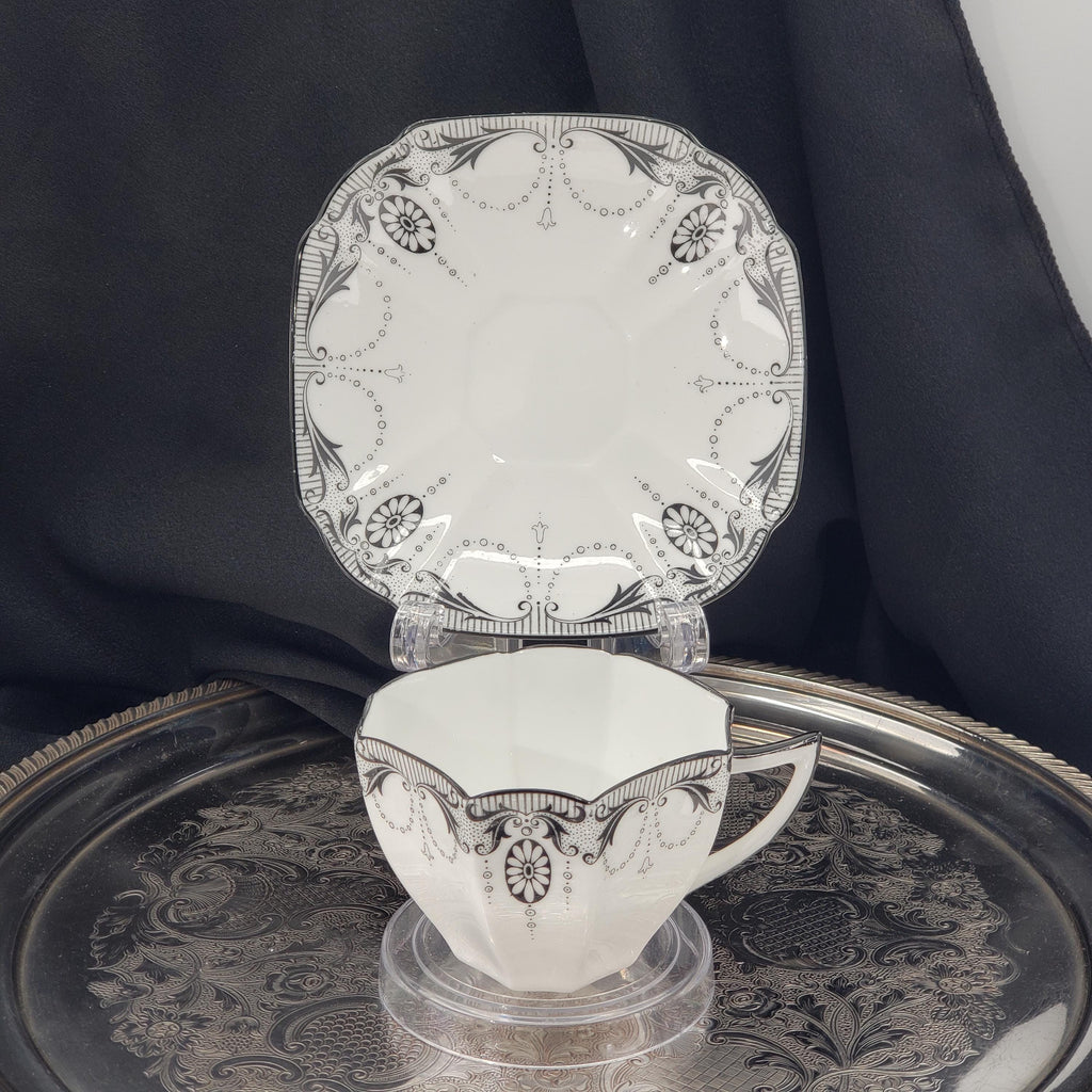 A white ceramic tea cup with an ornate black and white floral design sits on a silver tray, accompanied by a matching saucer.