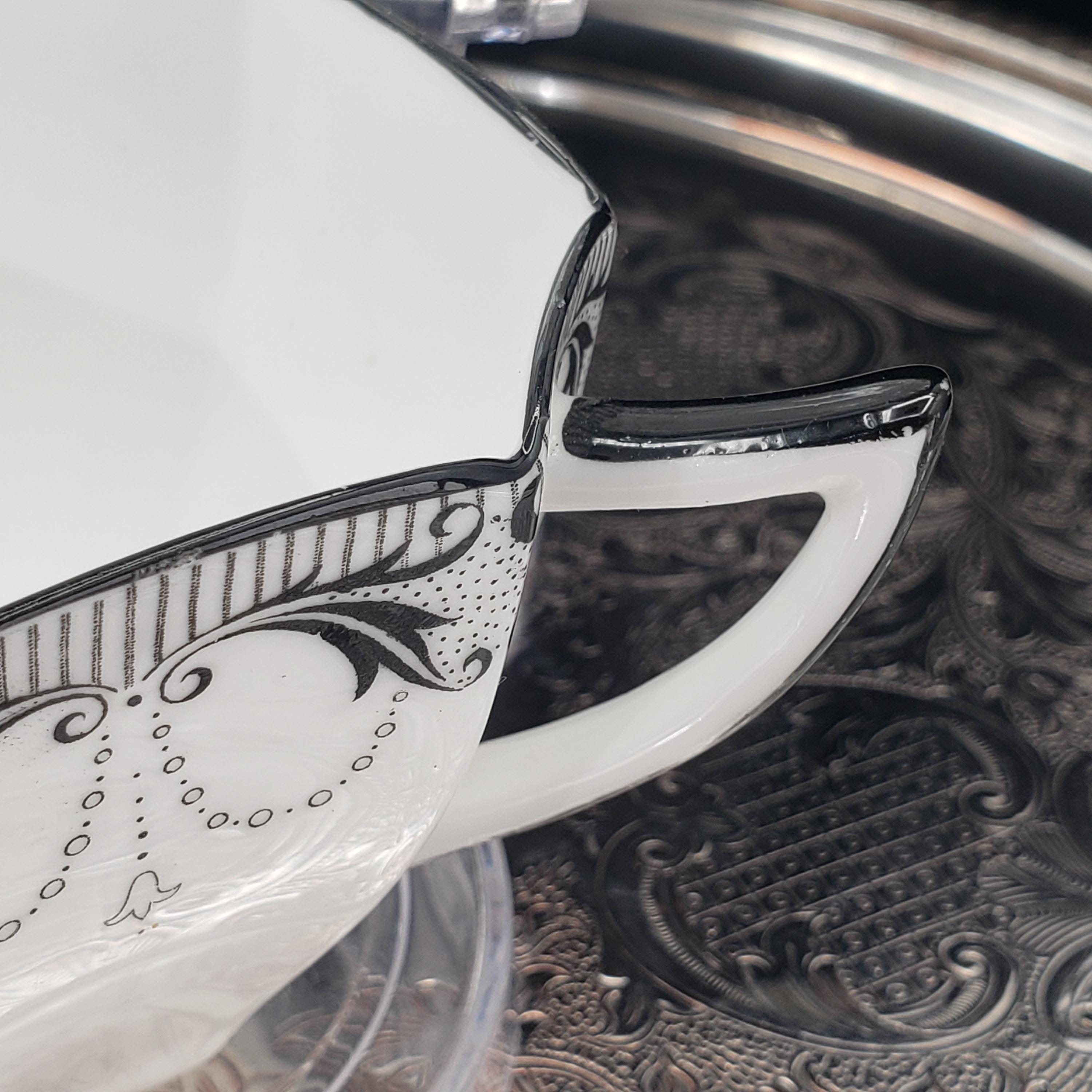 A close-up view of a white ceramic mug with a black and white floral design on its side, resting on a metal tray.
