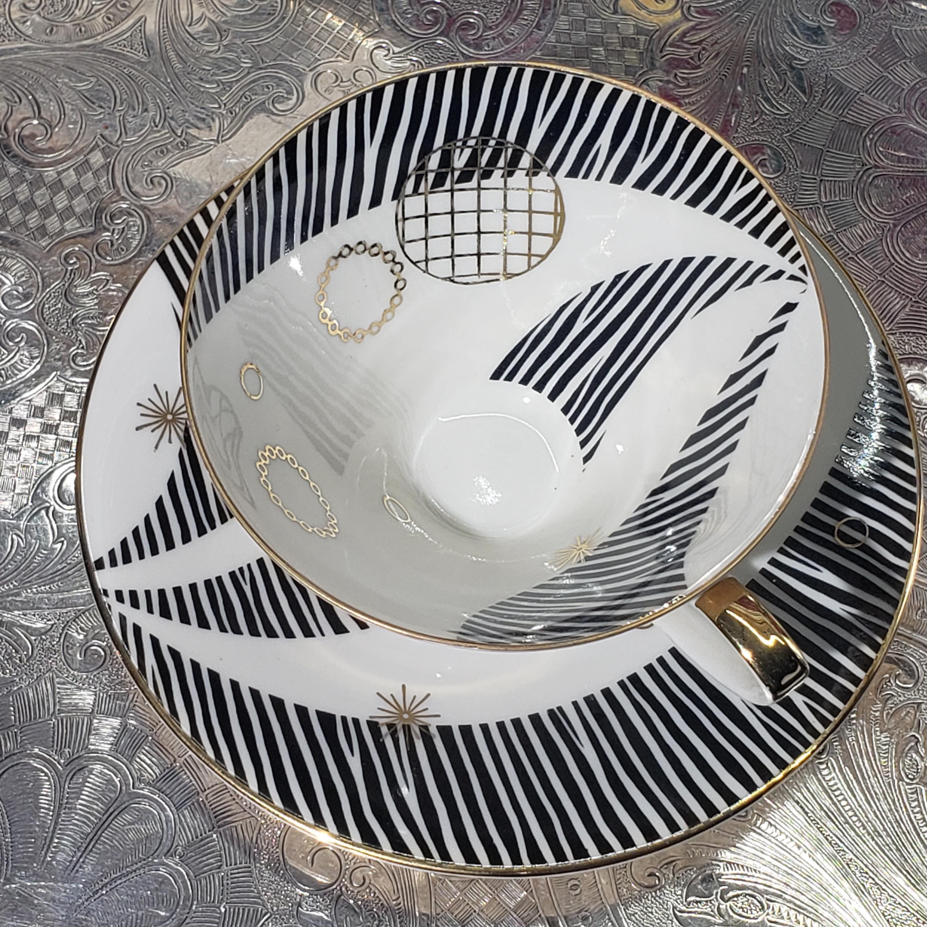 A white teacup with black and gold stripes and a gold handle, placed on a matching saucer with a similar design.