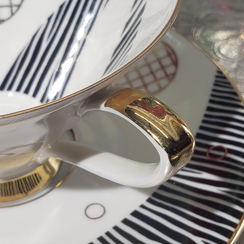 A close-up view of a white ceramic mug with a gold handle, resting on a white saucer with a black and white striped pattern.