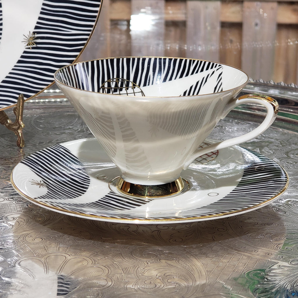 A white teacup with a black and white striped pattern sits on a matching saucer, placed on a patterned tablecloth.