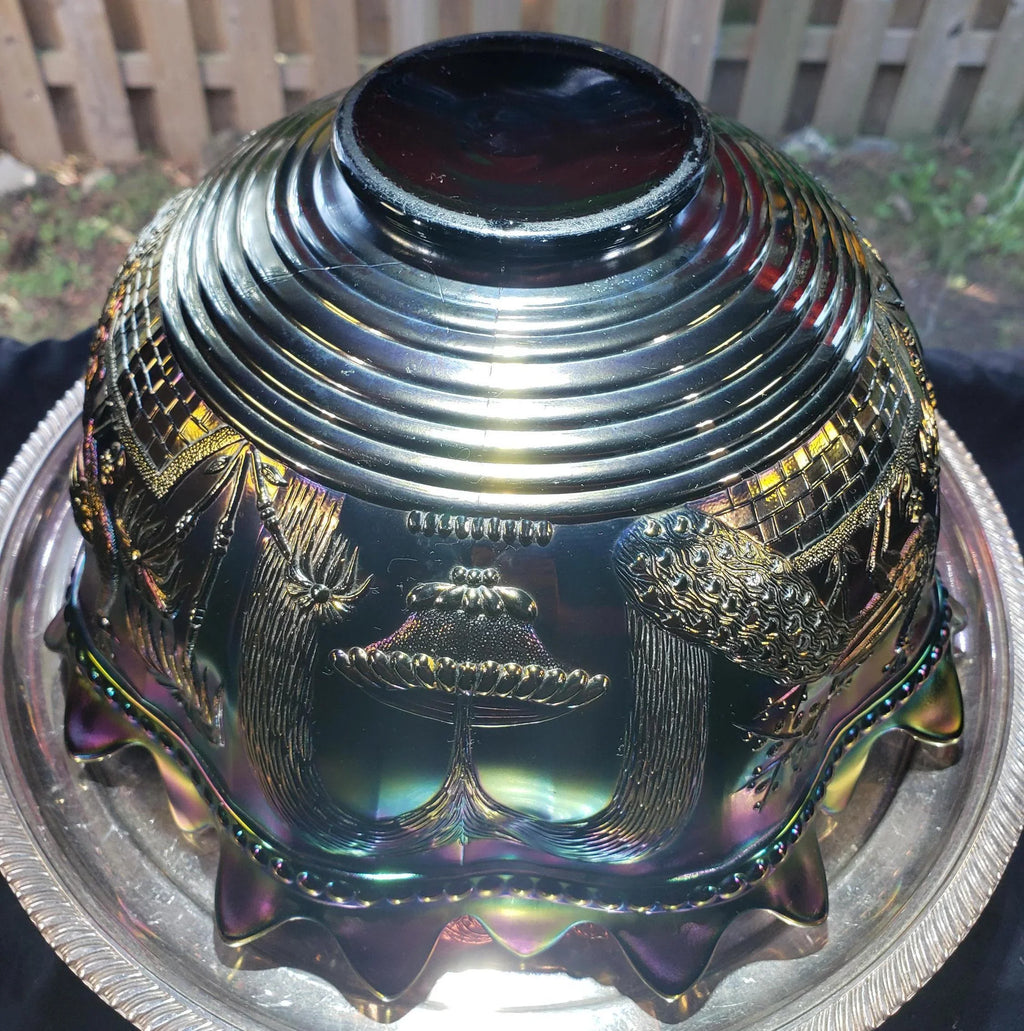 a decorative bowl with intricate patterns and designs, including a central floral motif and a bird-like figure. The bowl is placed on a metal tray, which is set against a wooden fence background.